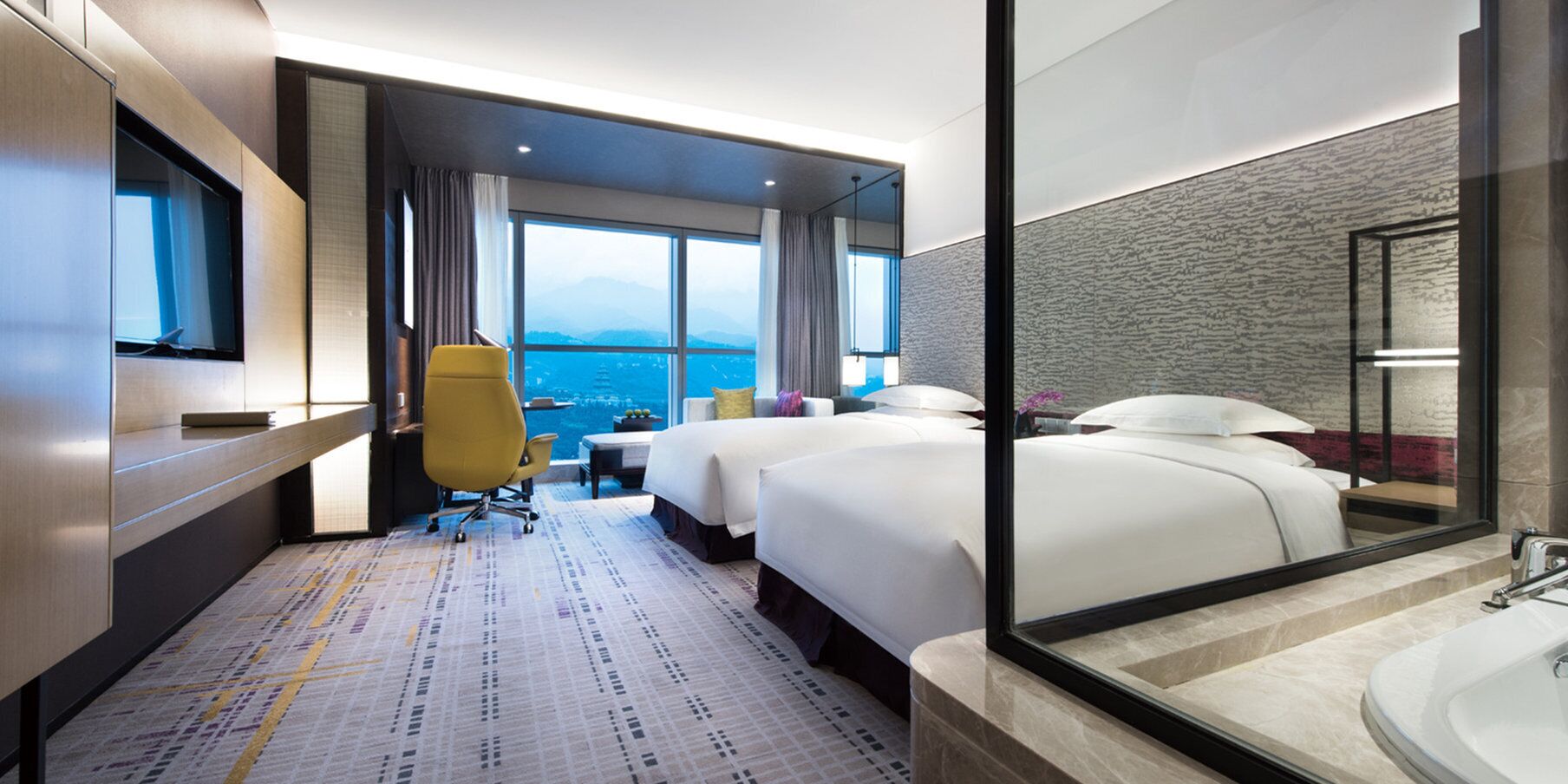 A hotel room with two beds, a desk, a chair, a large window with a view, and a bathroom with a mirror and sink.