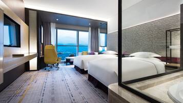 A hotel room with two beds, a desk, a chair, a large window with a view, and a bathroom with a mirror and sink.