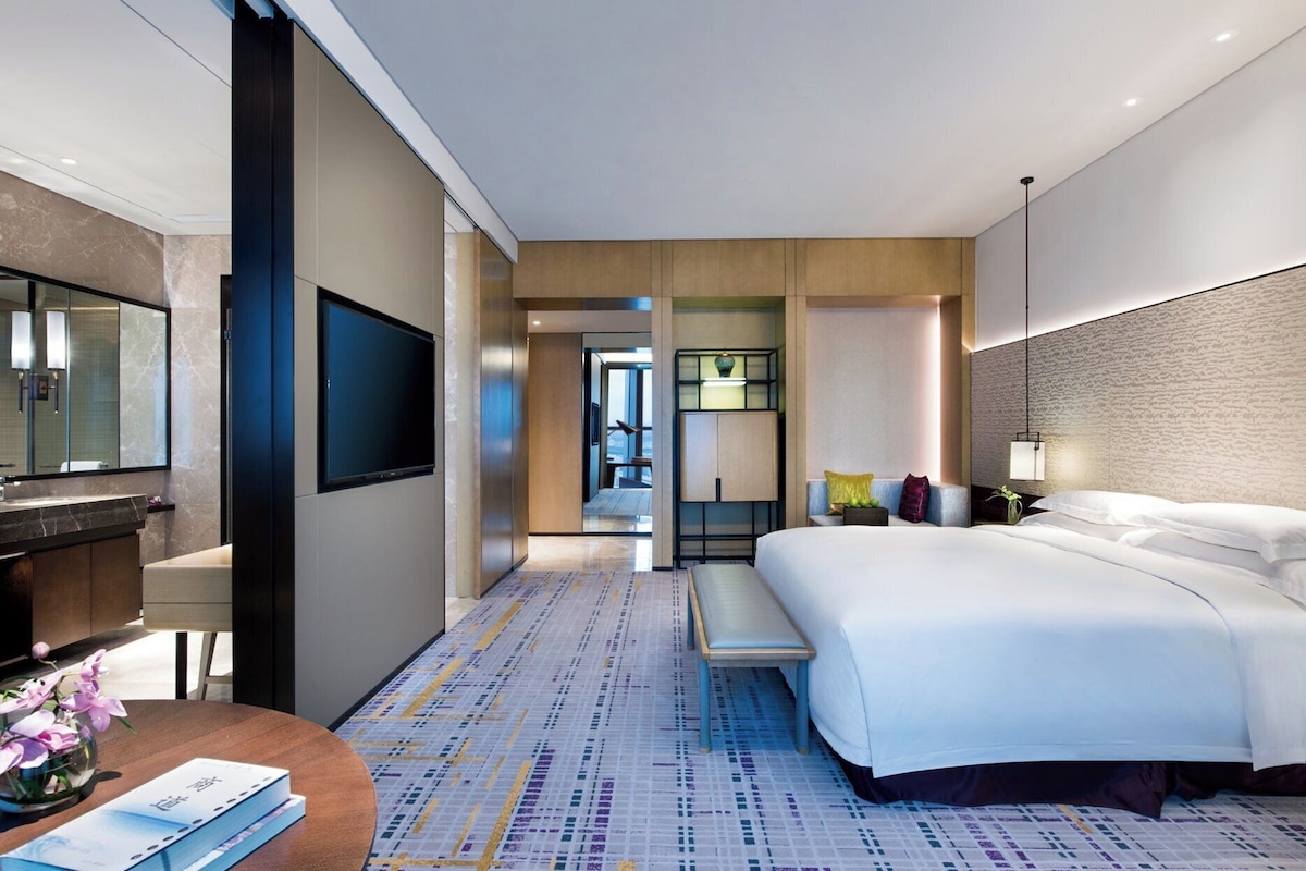 A modern hotel room with a large bed, a flat-screen TV, a desk with a chair, and a small table with a vase of flowers.