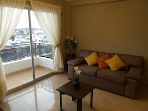 Ein Wohnzimmer mit braunem Sofa, einem kleinen Tisch und Blick auf eine Stadtlandschaft durch ein großes Fenster mit weißen Vorhängen.
