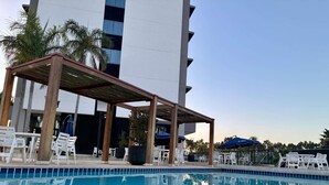 A pool area with a wooden pergola, white lounge chairs, and umbrellas. - Ibis Styles Taubate (Taubate)