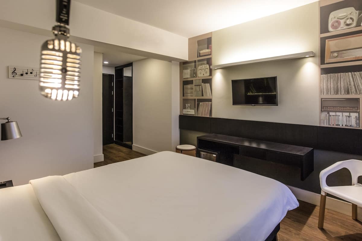 Une chambre d’hôtel moderne équipée d’un grand lit, d’un bureau, d’une chaise, d’un téléviseur fixé au mur et d’une bibliothèque contenant divers objets.