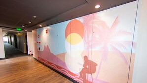 A hallway with a wooden floor and a wall mural depicting a sunset scene with palm trees and a silhouette of a person. - Ibis Styles Taubate (Taubate)