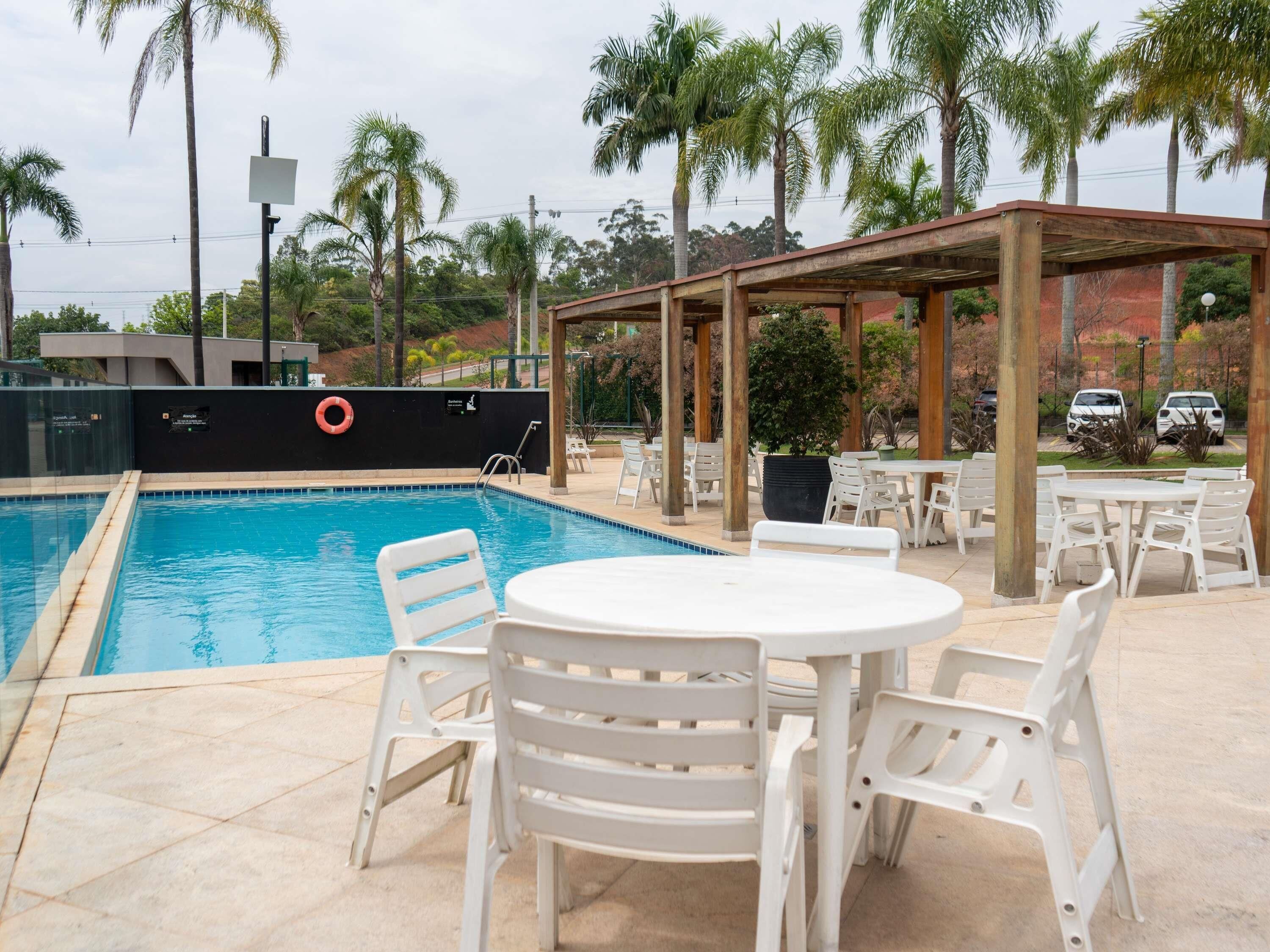 Una piscina con sillas y mesas blancas, rodeada de palmeras y una zona cubierta.