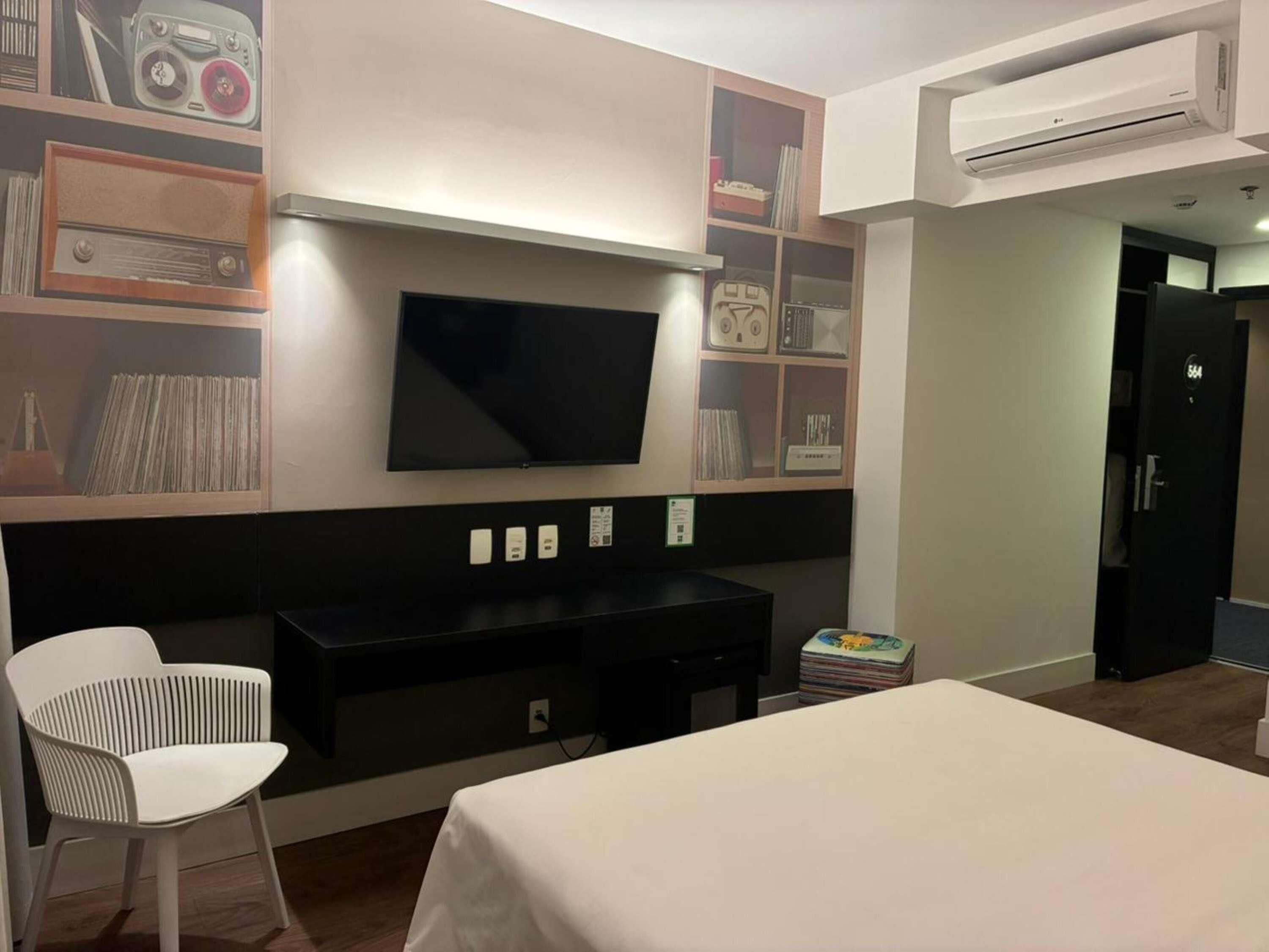 A hotel room with a bed, a TV mounted on the wall, a desk, a chair, and an air conditioning unit.