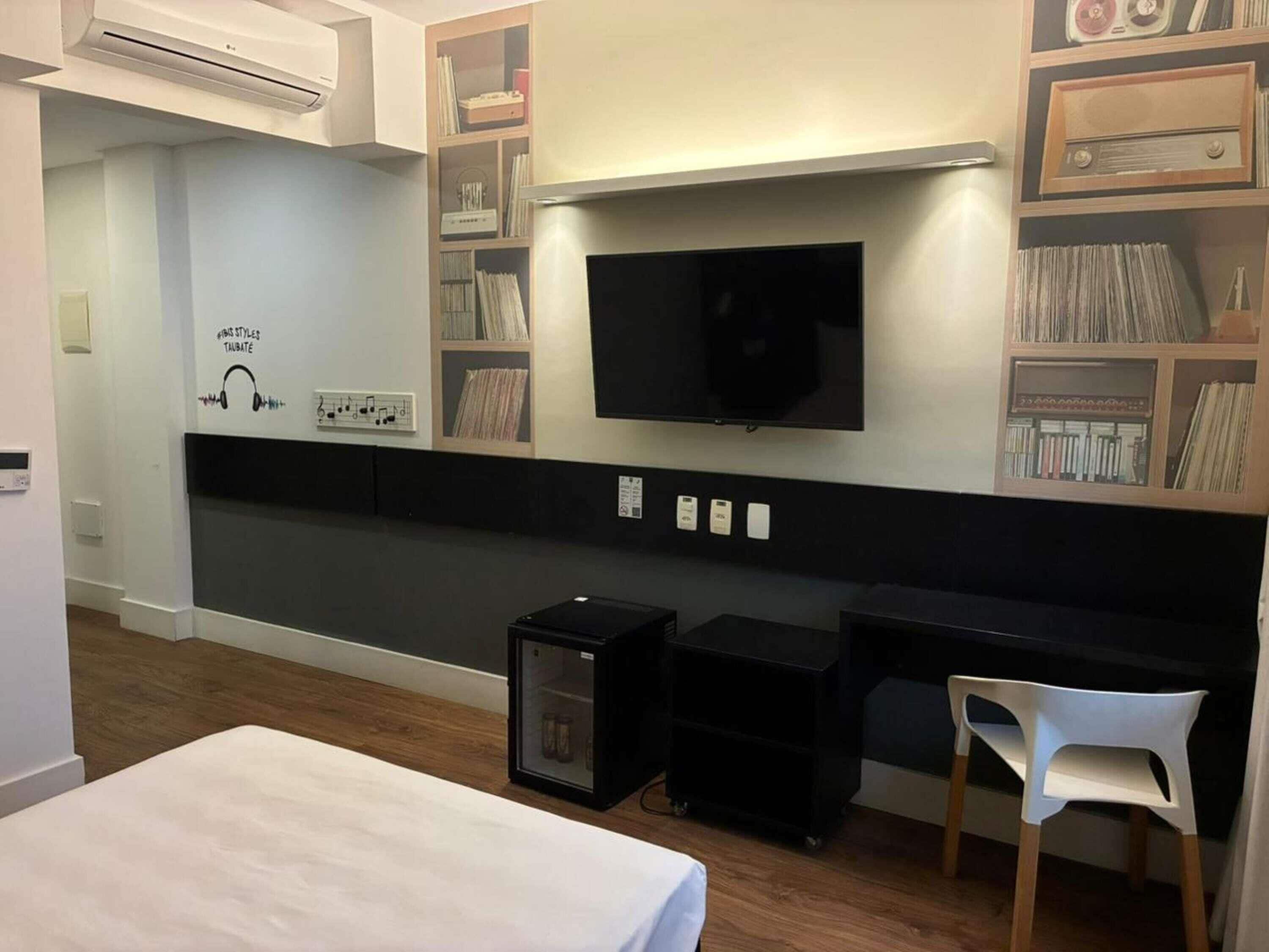 A modern hotel room with a flat-screen TV, built-in shelving with books and records, a small refrigerator, and a white chair.