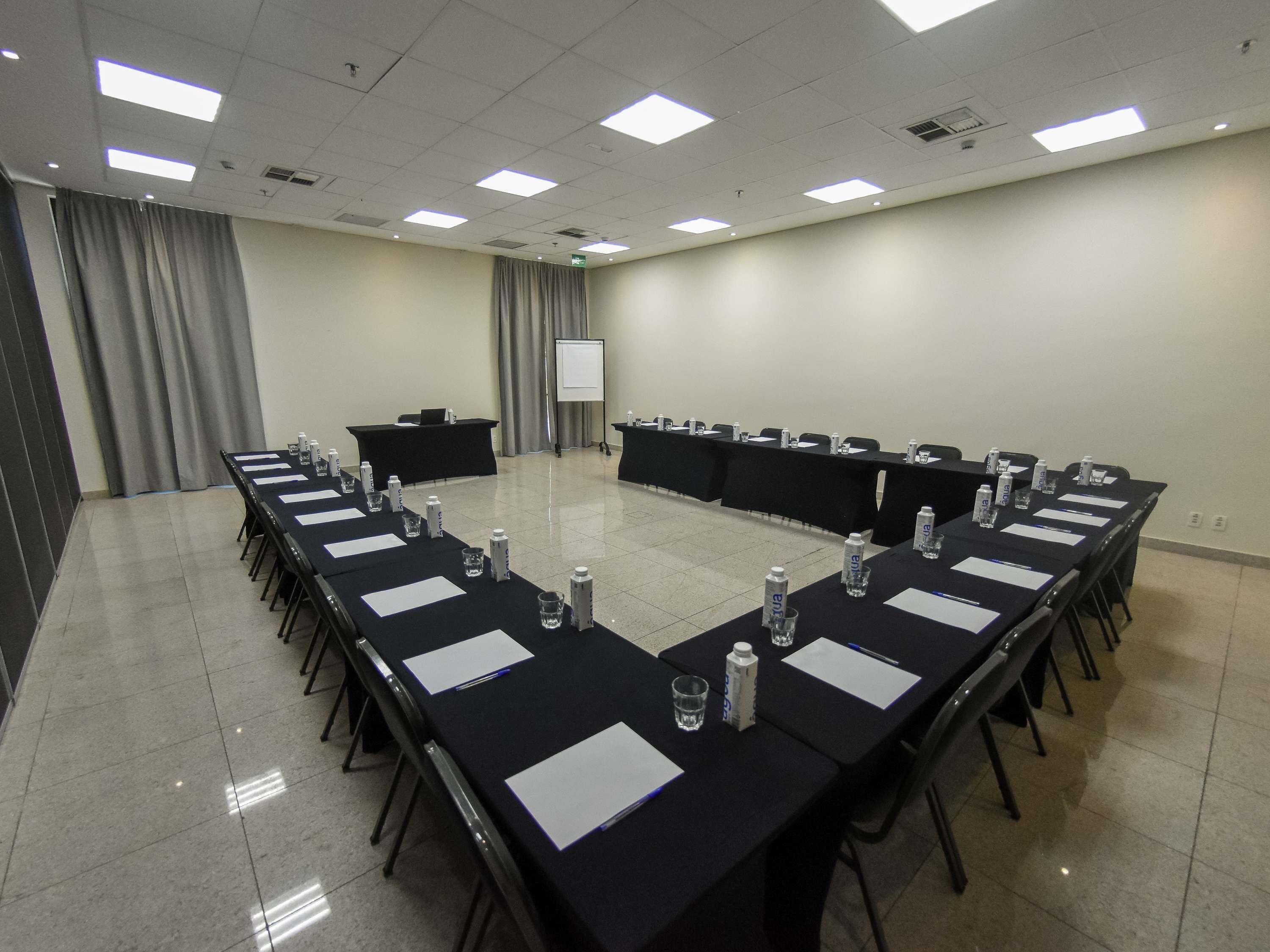 Un salón de conferencias con una mesa larga, sillas, un atril, una pantalla de proyector y botellas de agua.