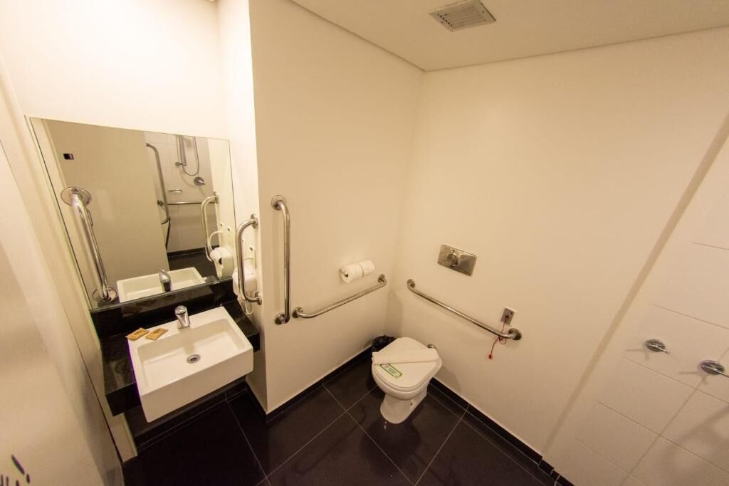 A small, modern bathroom with a white sink, mirror, toilet, and handrails.