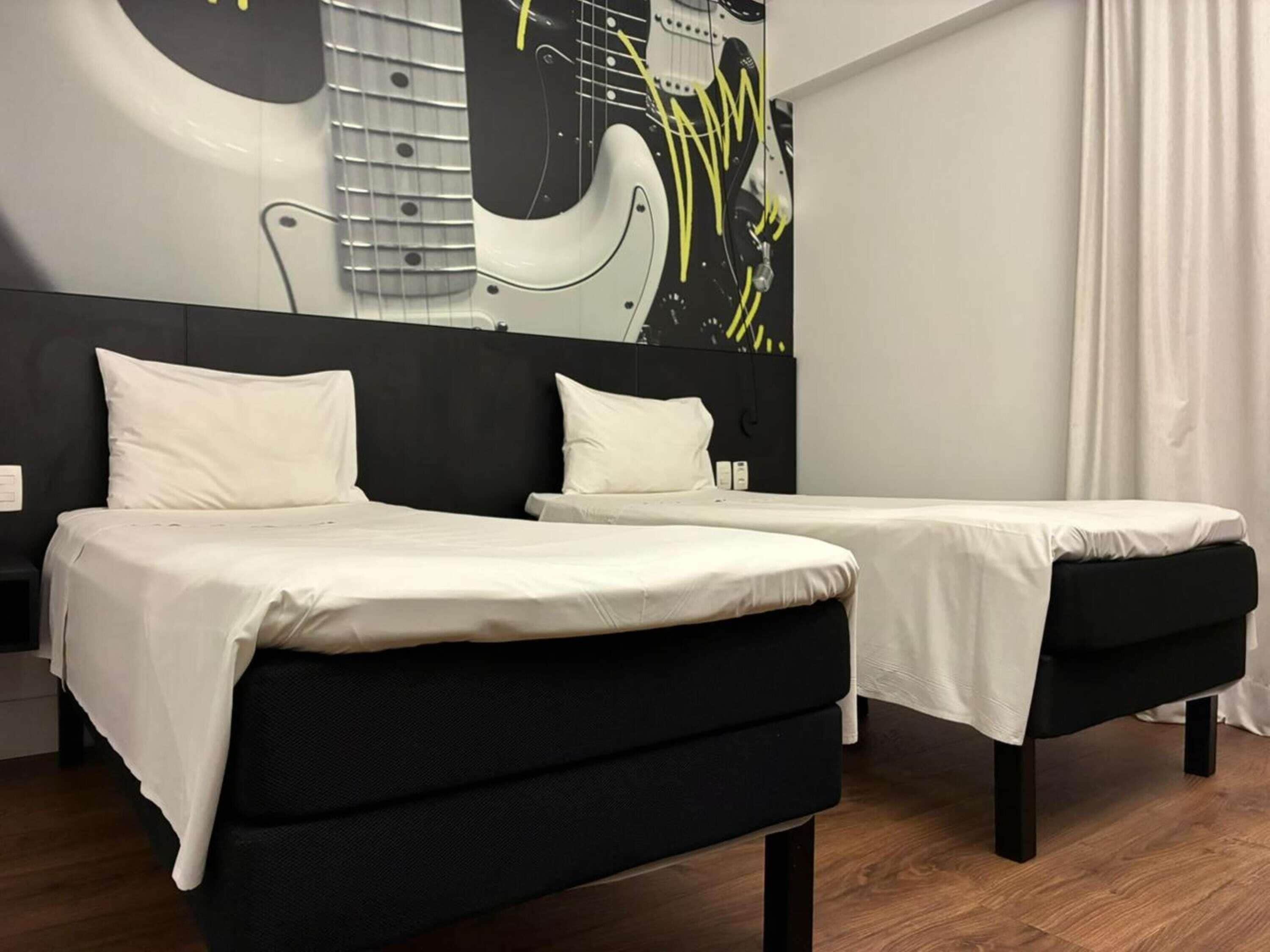 Una camera d'albergo con due letti, una testiera nera e un murale con un'immagine di una chitarra.