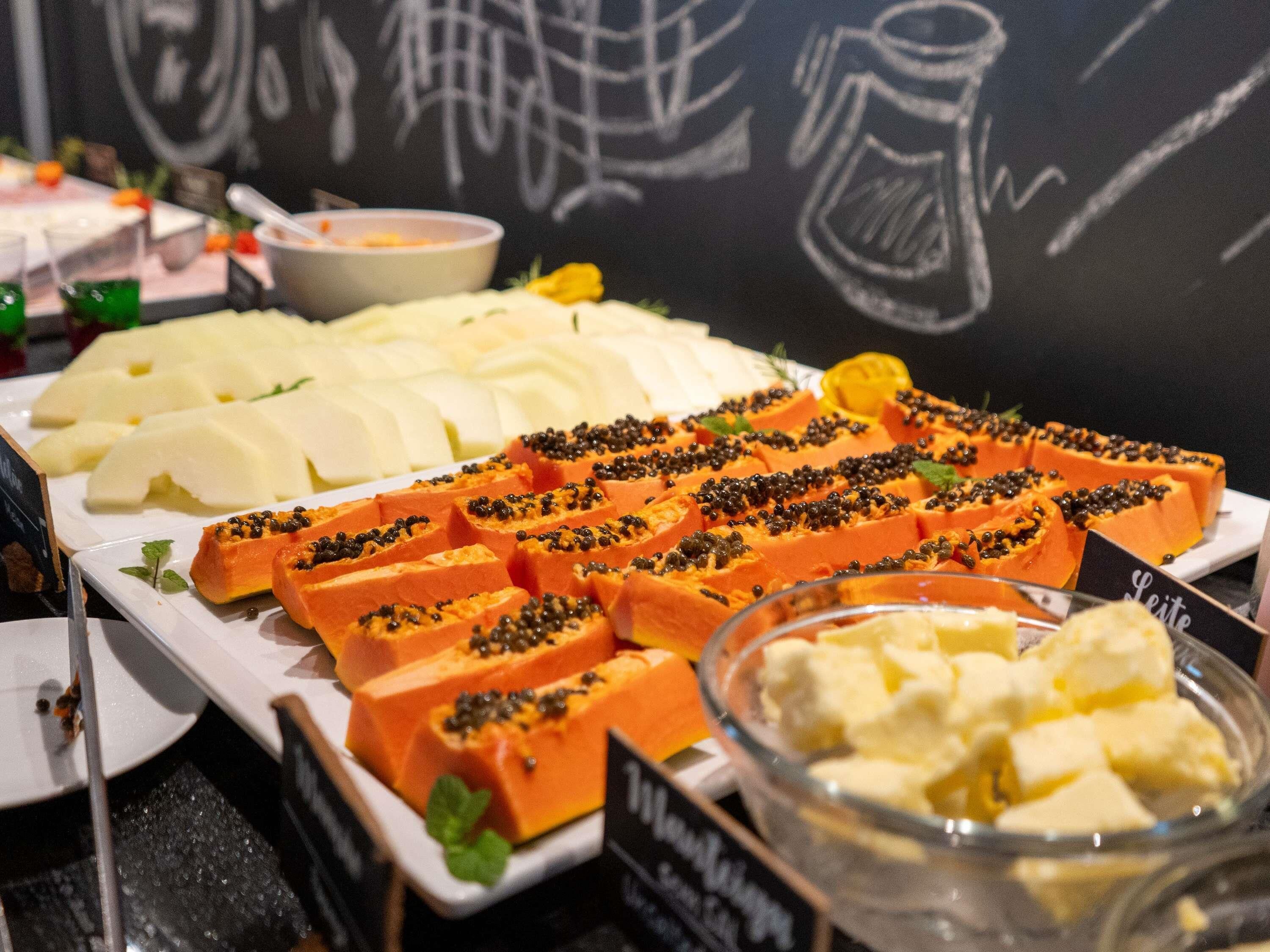 Un bufé con diversos alimentos, incluyendo frutas blancas y naranjas en rodajas, un recipiente con comida amarilla y una pizarra con dibujos.