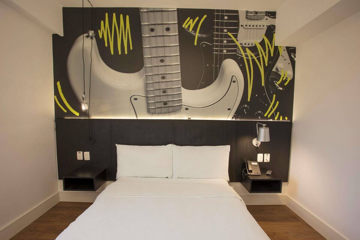 Une chambre d’hôtel moderne avec un grand lit, une tête de lit ornée d’une œuvre d’art abstraite, des lampes de chevet et un téléphone fixé au mur.
