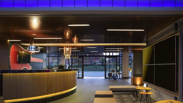 A modern reception area with a curved counter, a red guitar sculpture, and a drum set in the background. There are seating areas with patterned floor tiles and tables. - Ibis Styles Taubate (Taubate)