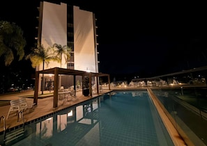 Un área de piscina nocturna con sillones de descanso y una zona de estar cubierta, rodeada por un edificio moderno con grandes ventanales.