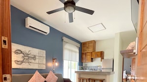 Sala de estar con sofá, mesa de centro, alfombra estampada, ventilador de techo y zona de cocina con armarios y ventana.