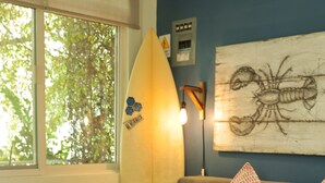 A room with a sofa, a wooden coffee table with a vase of flowers, a surfboard decoration, and a wall with a lobster artwork. - PUNTO MITA ECO SURF SUITES (Punta de Mita)