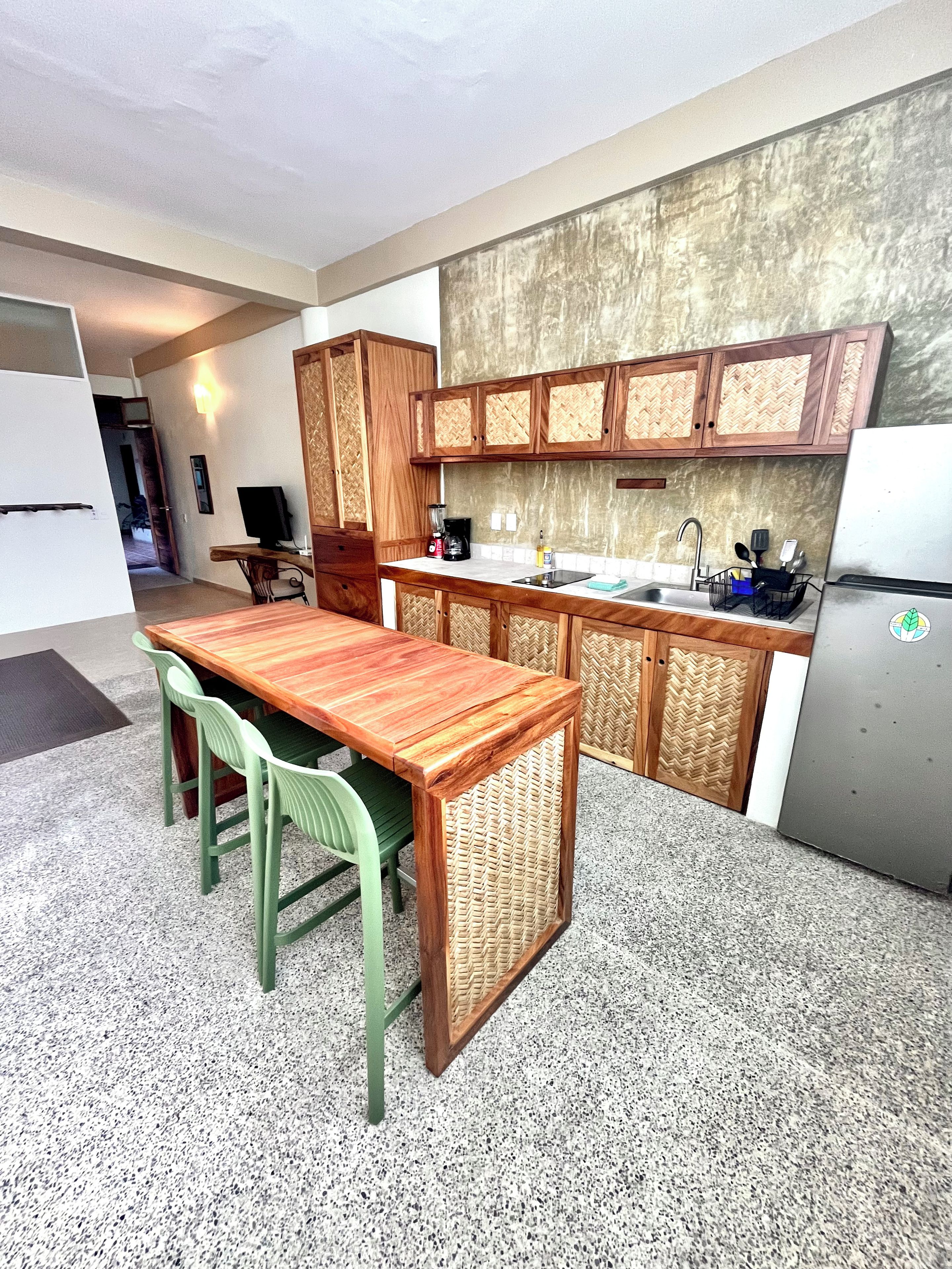 Una cocina compacta con una mesa de madera, sillas verdes, un armario empotrado con diseño en espiga, un refrigerador de acero inoxidable y una pequeña encimera con un fregadero y diversos utensilios de cocina.