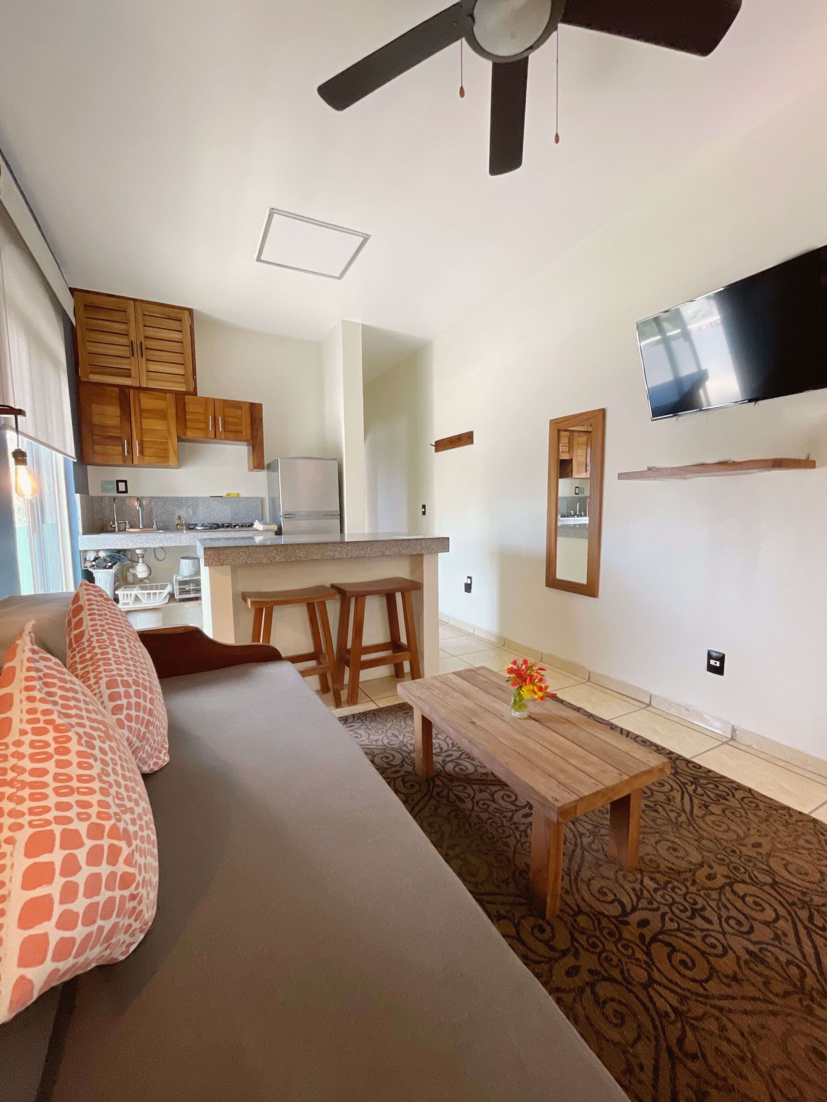 A modern kitchen with wooden cabinets, a dining area with a table and chairs, a ceiling fan, and a mounted TV.
