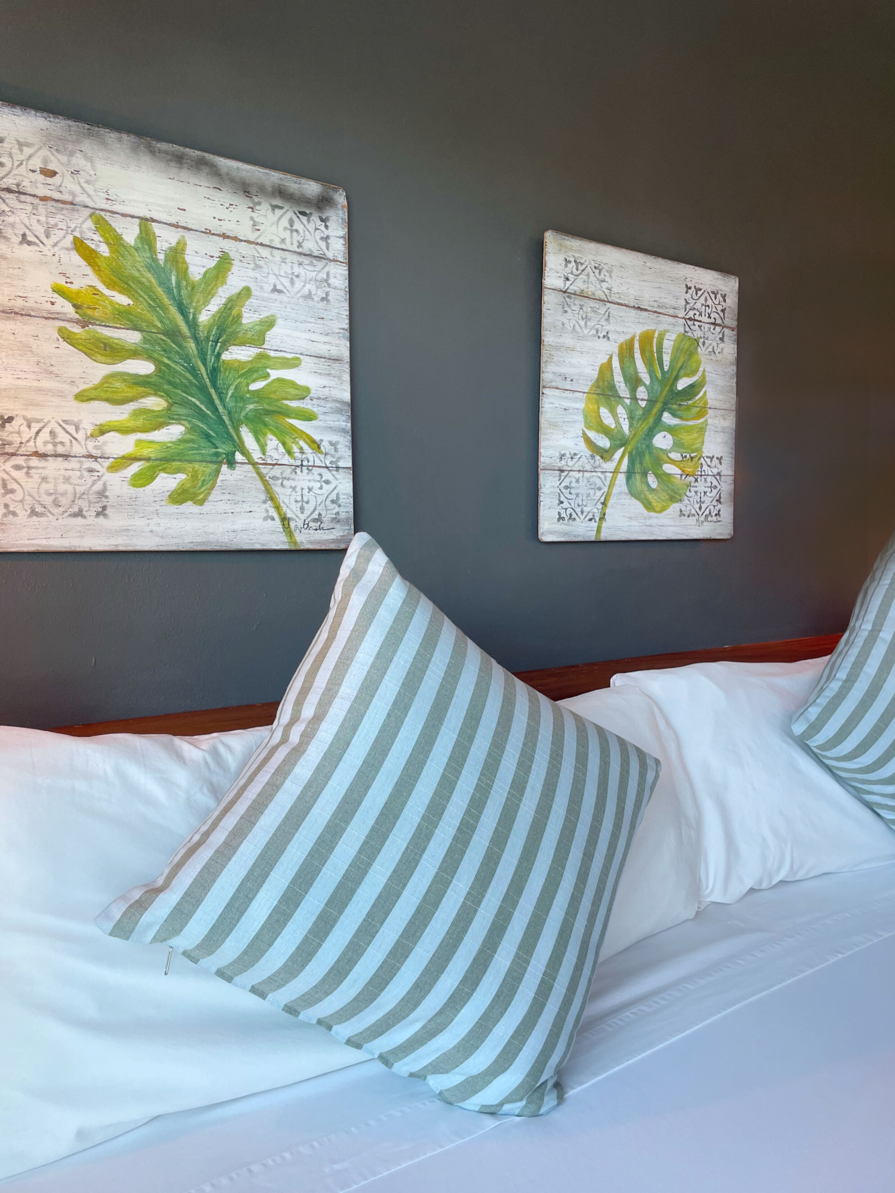 Ein Bett mit weißer Bettwäsche und gestreifter Kopfkissen, zwei an der Wand befestigte Kunstwerke mit Blättern und ein Holz-Kopfbrett.