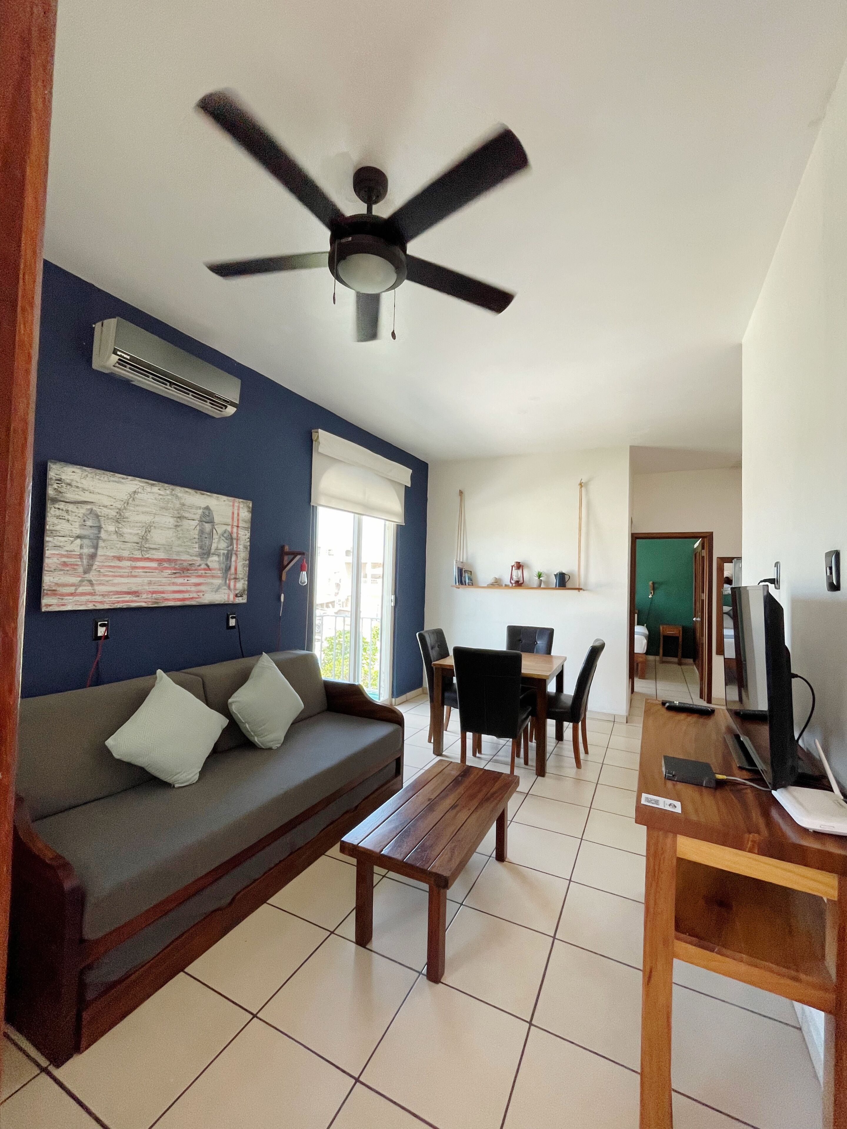 A living room with a sofa, a wooden bench, a ceiling fan, an air conditioner, a dining table with chairs, and a television.