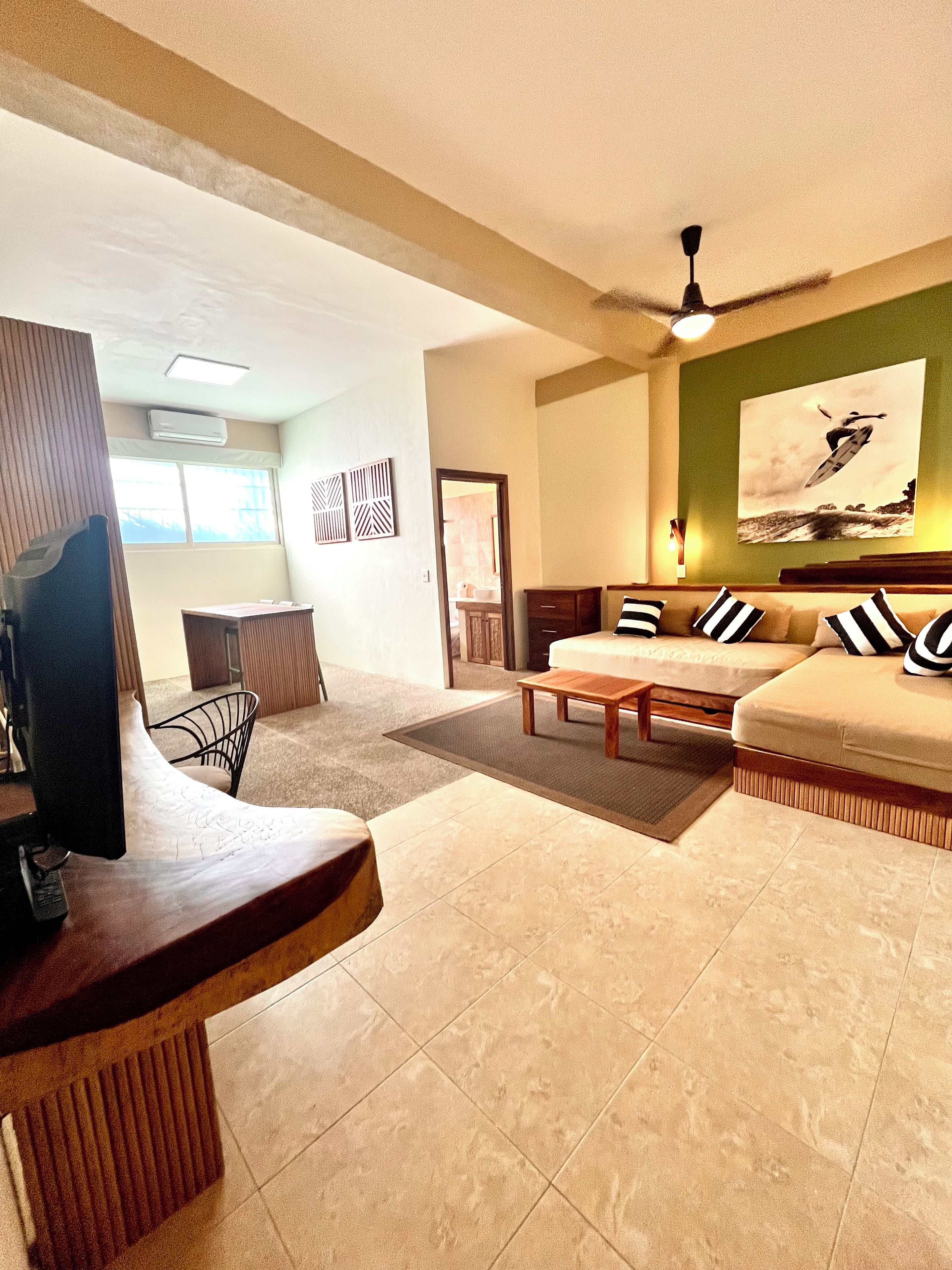 A living room with a sofa, coffee table, and a television. There is a ceiling fan, a painting on the wall, and a window.