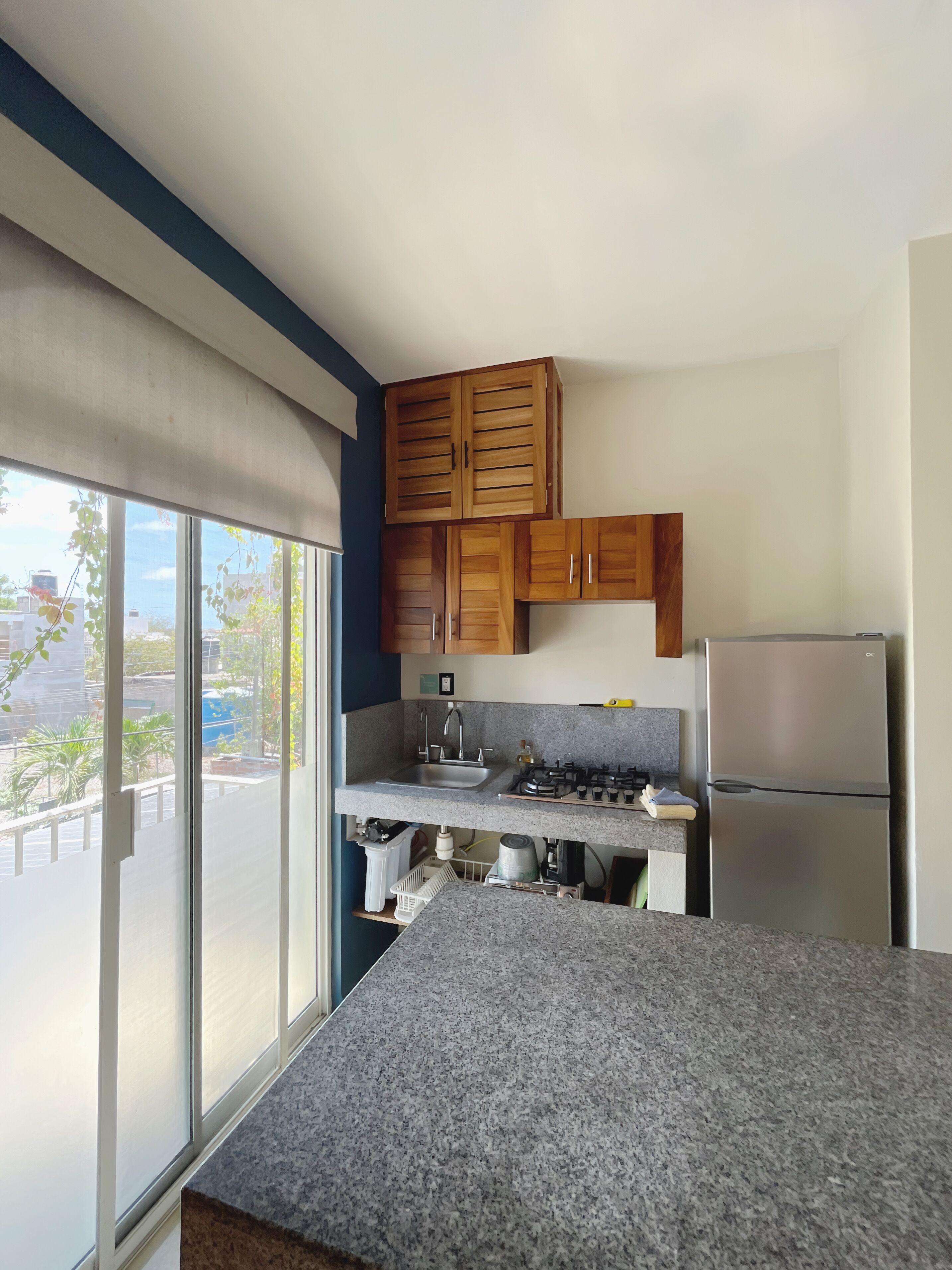 Una cocina con encimera de granito, armarios de madera, refrigerador de acero inoxidable y ventana con vista a edificios y vegetación.