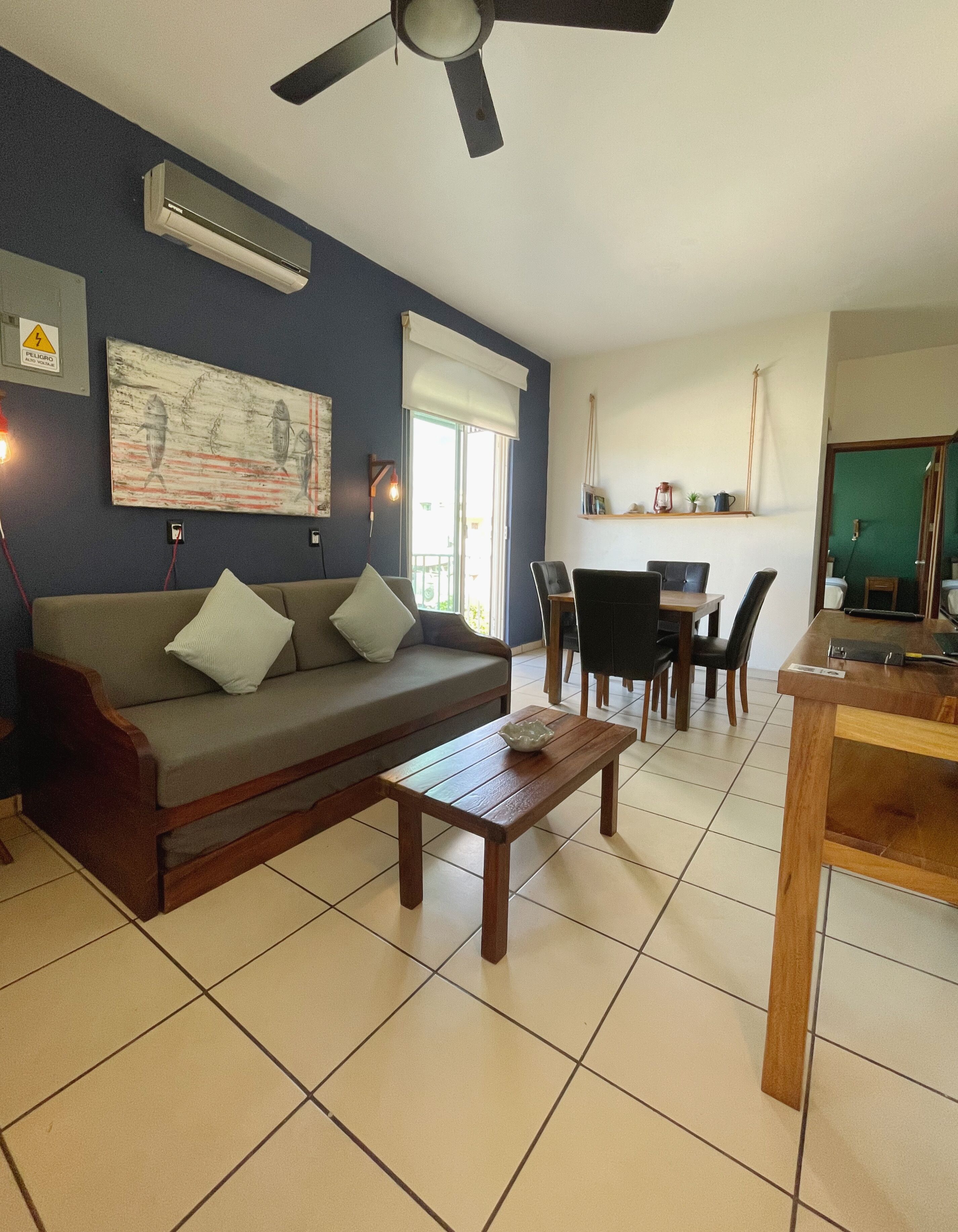Een woonkamer met een bank, salontafel, eettafel, stoelen, een plafondventilator, airconditioning, kunstwerken en een raam met jaloezieën.