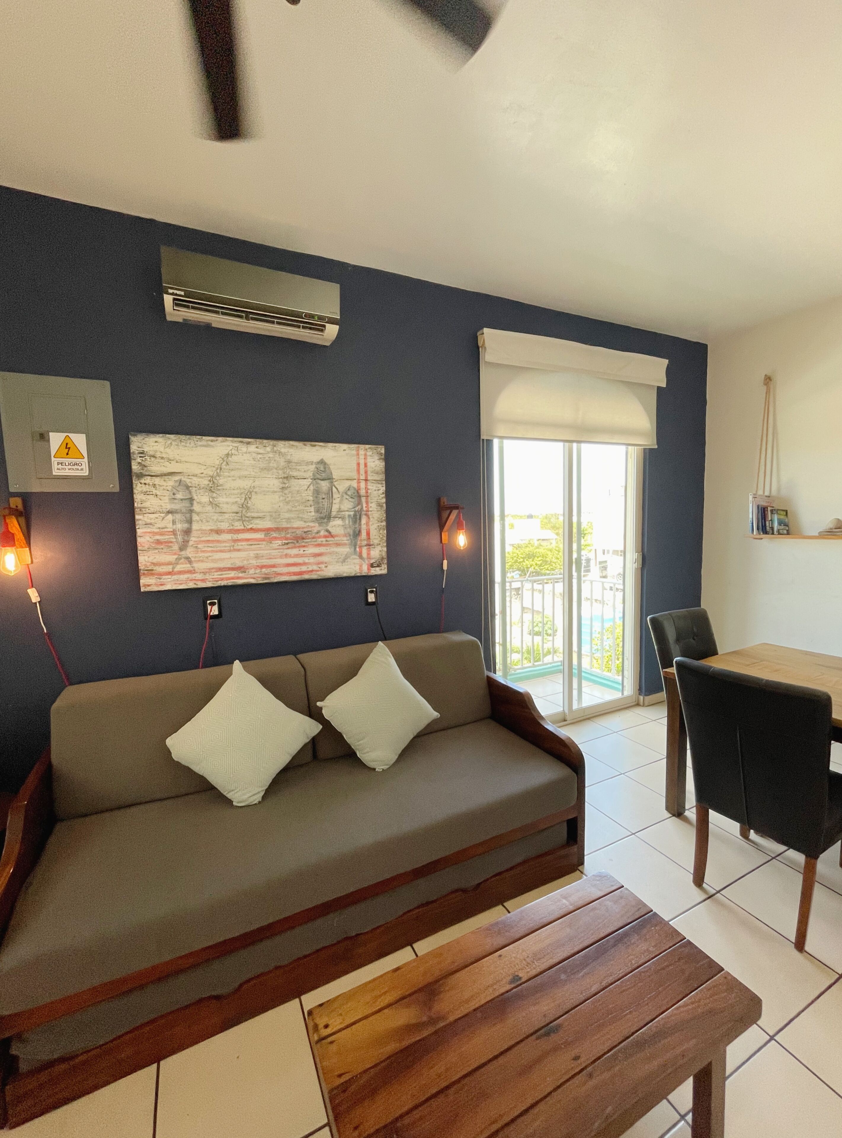 A living room with a sofa, a wooden coffee table, a dining table with chairs, a painting on the wall, an air conditioning unit, and a window with a view.