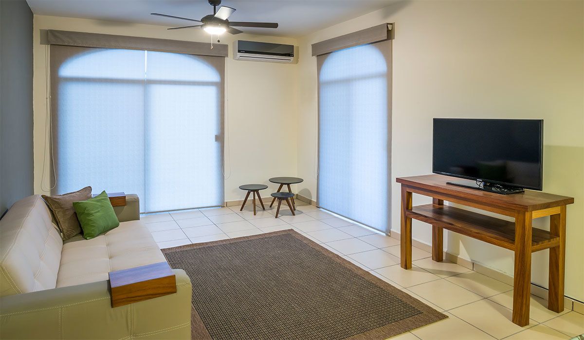 Un salon avec un canapé, un téléviseur posé sur un meuble en bois, un ventilateur de plafond et deux petites tables.