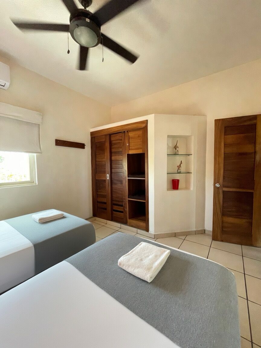 Une chambre avec deux lits, un ventilateur de plafond, des armoires en bois, une petite étagère avec des objets décoratifs et une fenêtre avec des stores.