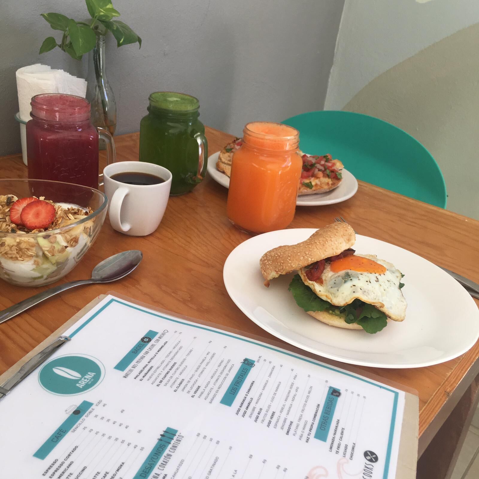 Una mesa de desayuno con un menú, un brebaje verde, un batido, un plato con un sándwich, un tazón de cereal y una taza de café.