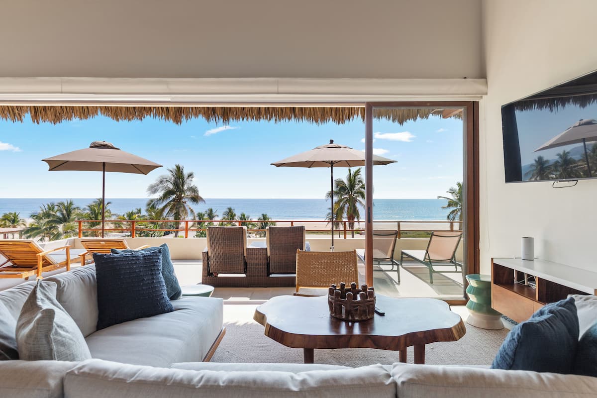 Una sala de estar moderna con un amplio ventanal con vista a la playa, amoblada con un sofá, sillones y una mesa de centro.