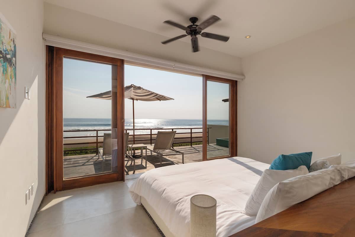 Un dormitorio con una cama grande, un ventilador de techo y vistas a la playa a través de ventanas de gran altura.