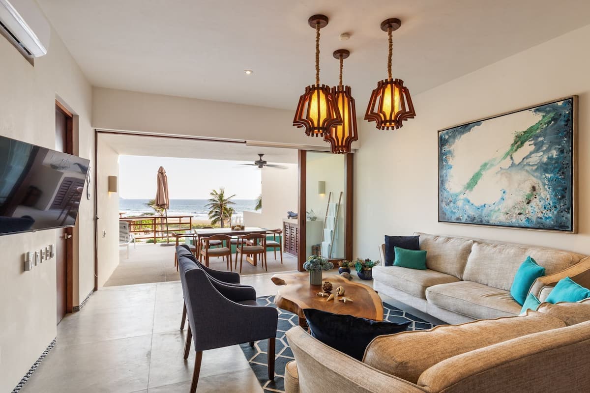Una sala de estar moderna con sofá, sillones, una mesa de centro y televisor. Hay una zona de comedor con una mesa y sillas, y vistas al océano.