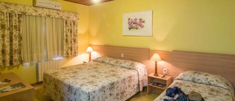 A hotel room with two beds, a wooden ceiling, an air conditioning unit, a window with curtains, a bedside table with a lamp, and a wall-mounted picture.
