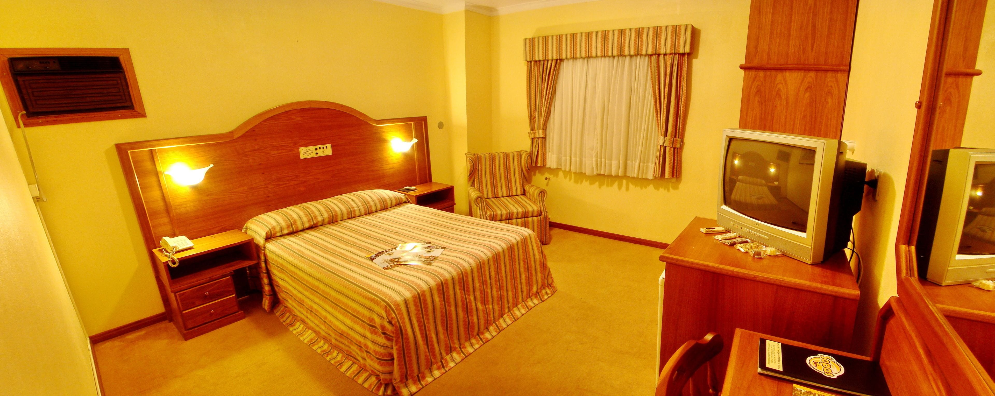 Een hotelkamer met een bed, nachtkastjes, een televisie, een stoel en een raam met gordijnen.