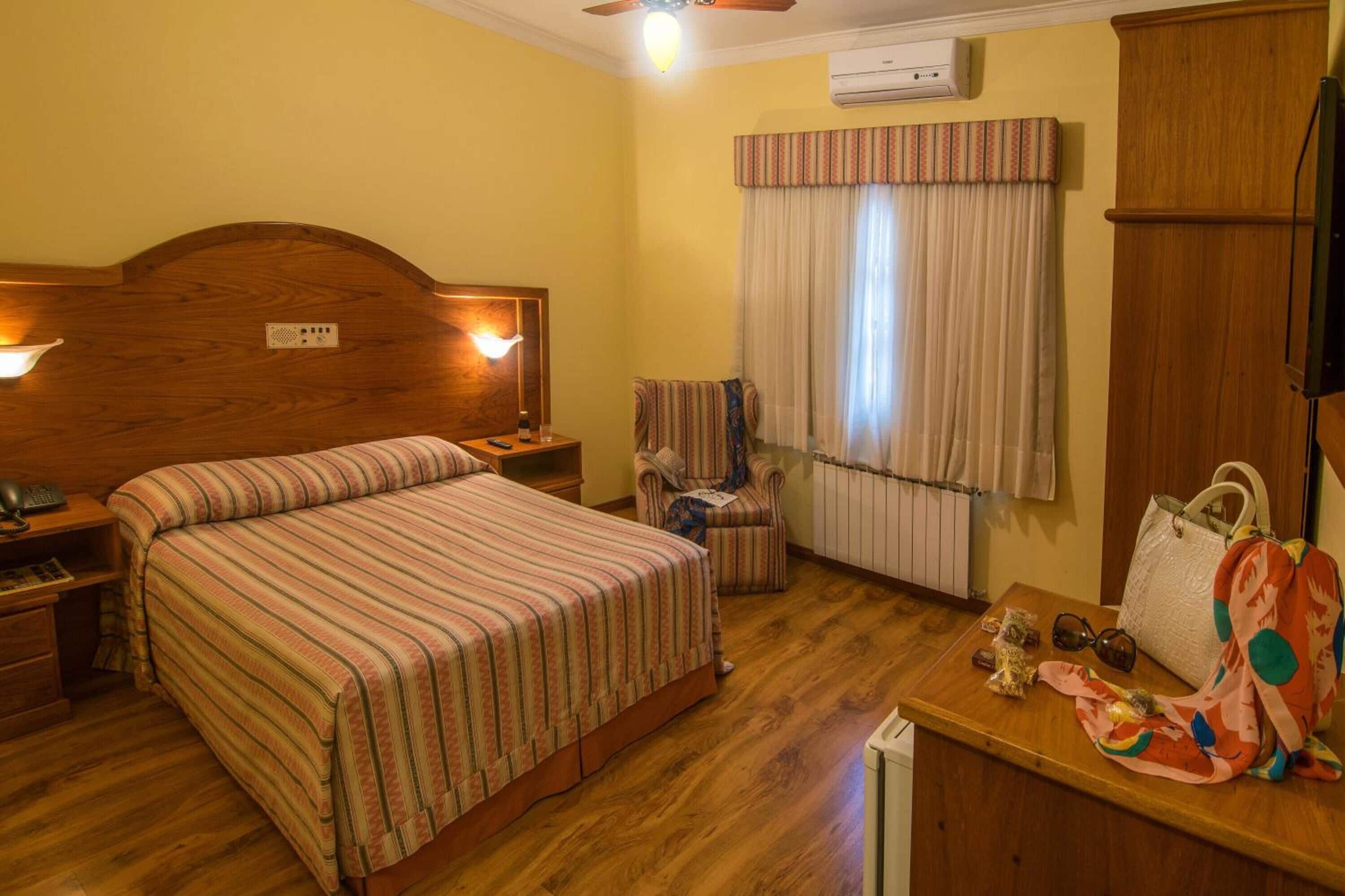 A hotel room with a wooden bed, two bedside lamps, a chair, a small table with personal items, and a window with curtains.