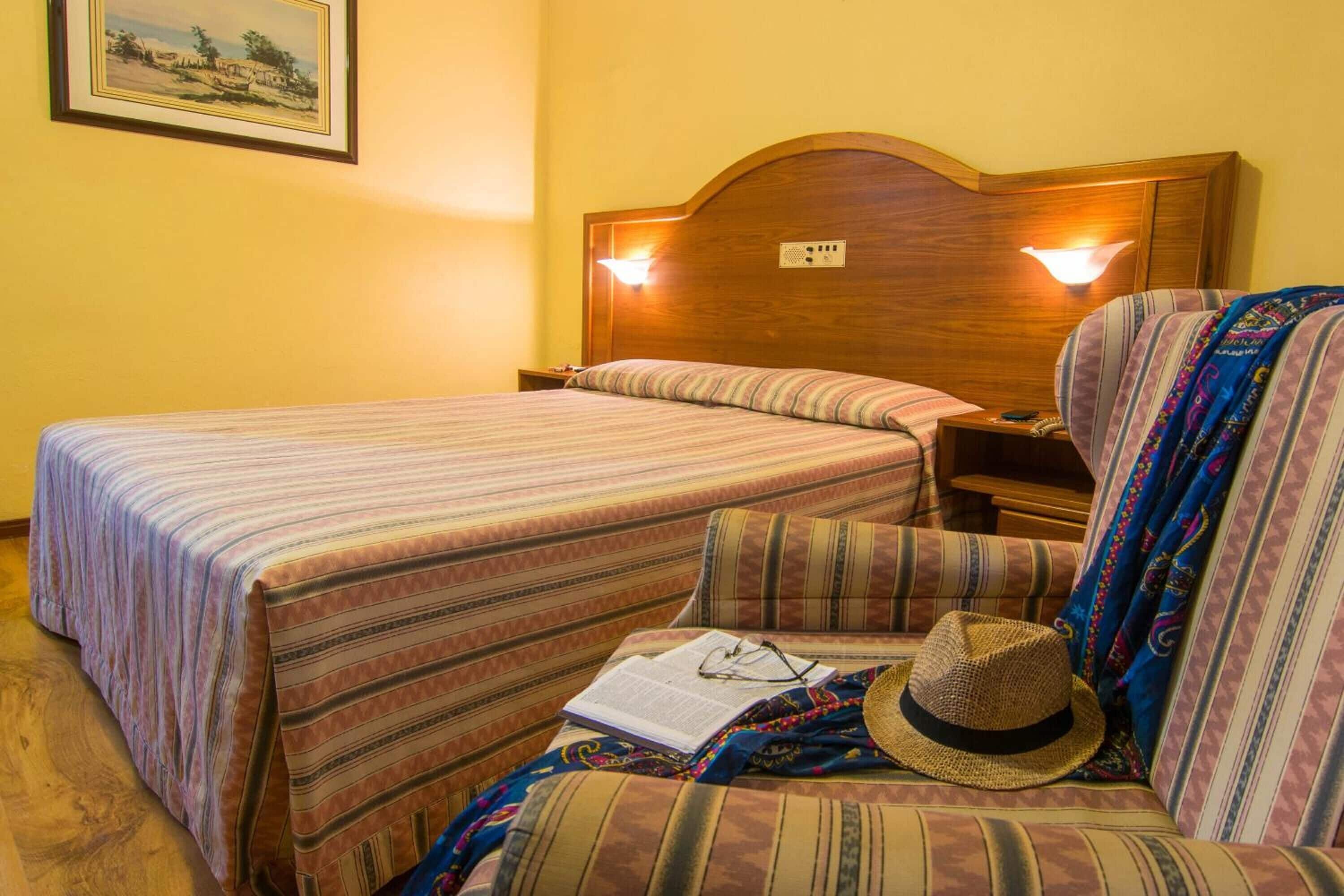 Habitación de hotel con dos camas, una mesita de noche, una lámpara, un sombrero y un libro sobre la cama.
