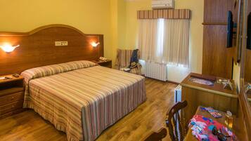 Habitación de hotel con una cama grande, cabecera de madera, un escritorio con una silla, una mesita con objetos y una ventana con cortinas.