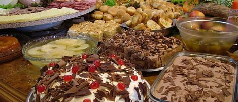 A buffet table with various dishes including a large cake with whipped cream and chocolate shavings, a bowl of fruit salad, and several platters of meats and cheeses.