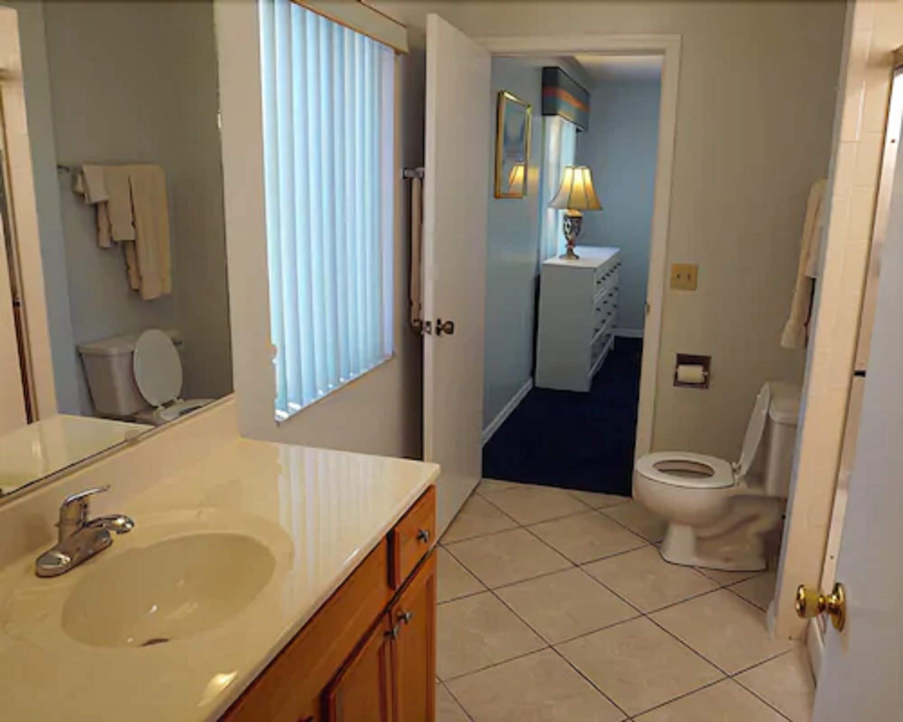 A bathroom with a sink, mirror, toilet, and a window with blinds.