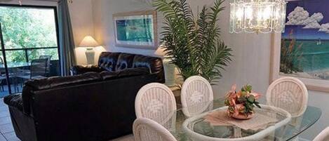 Un coin repas avec une table en verre entourée de chaises blanches, d’un canapé noir et d’une plante en pot.
