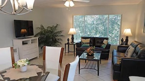 A living room with a dining table, chairs, a sofa, a TV, and a large window with blinds. - Vacation Villas (Titusville)