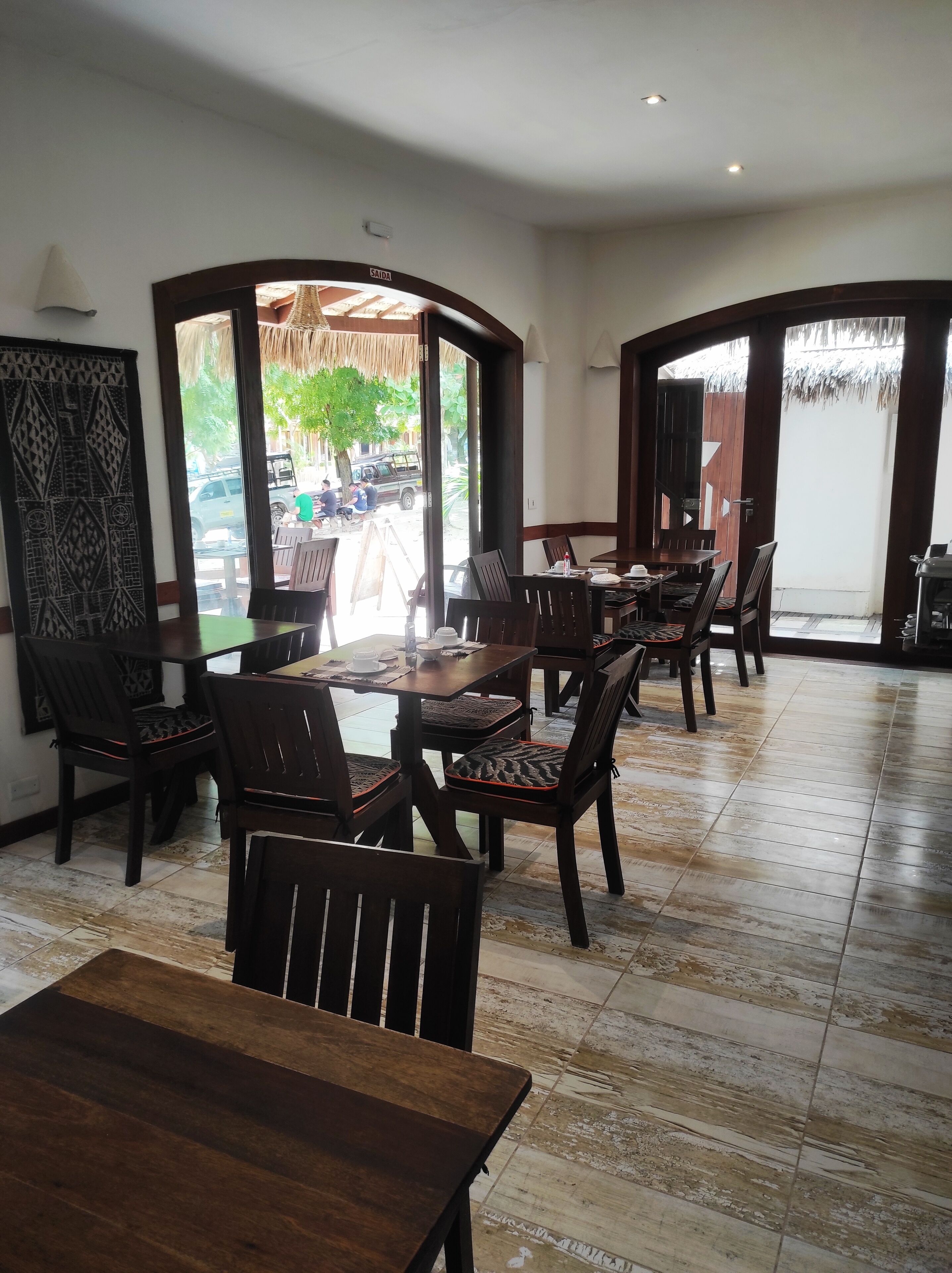 A dining area with wooden tables and chairs, a patterned wall, and a view of the outside.