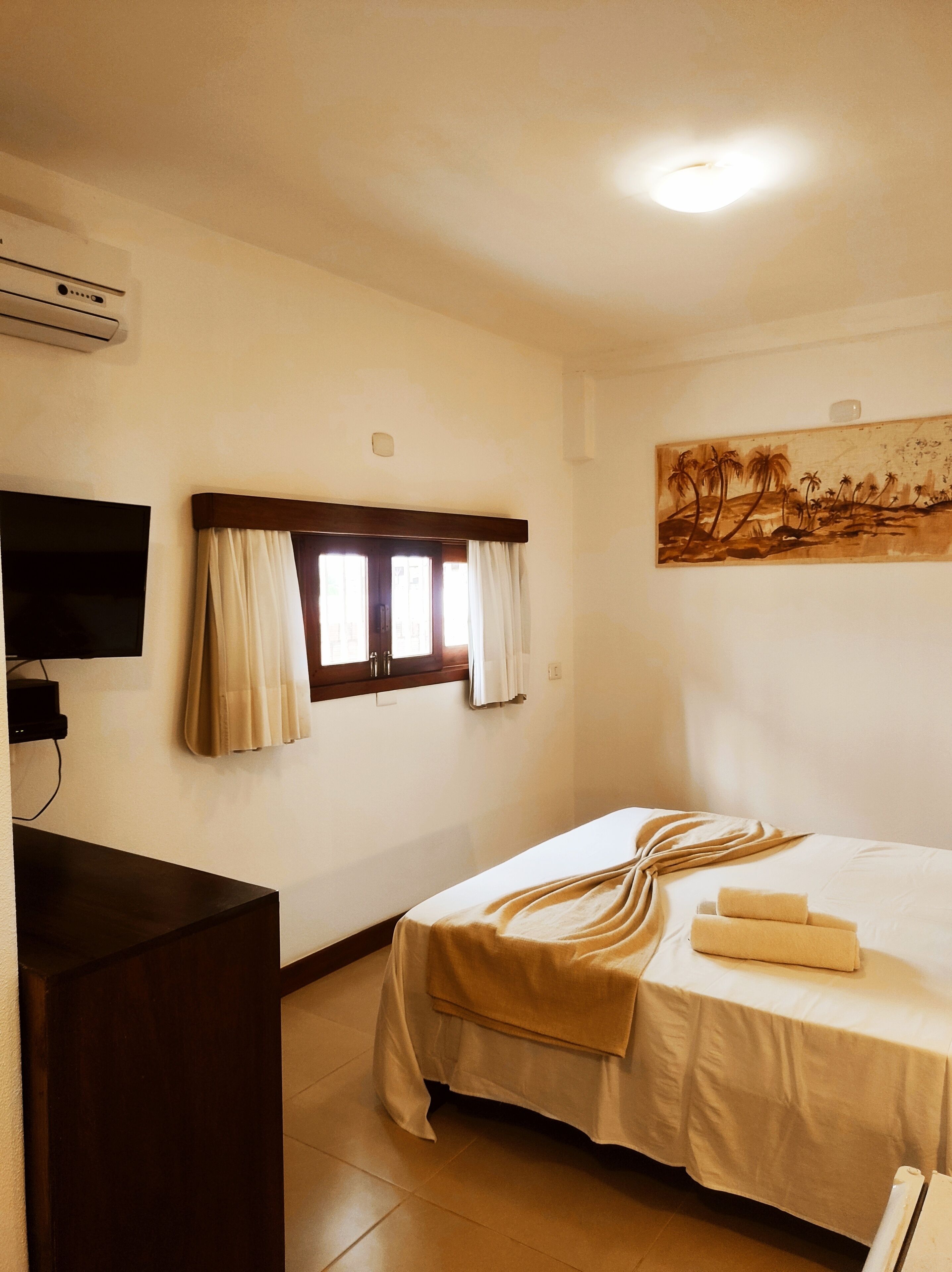 A hotel room with a bed, a flat-screen TV mounted on the wall, a window with curtains, and a painting on the wall.
