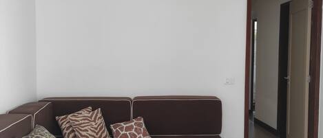 A living room with a brown sofa, patterned cushions, a standing fan, and a tiled floor.