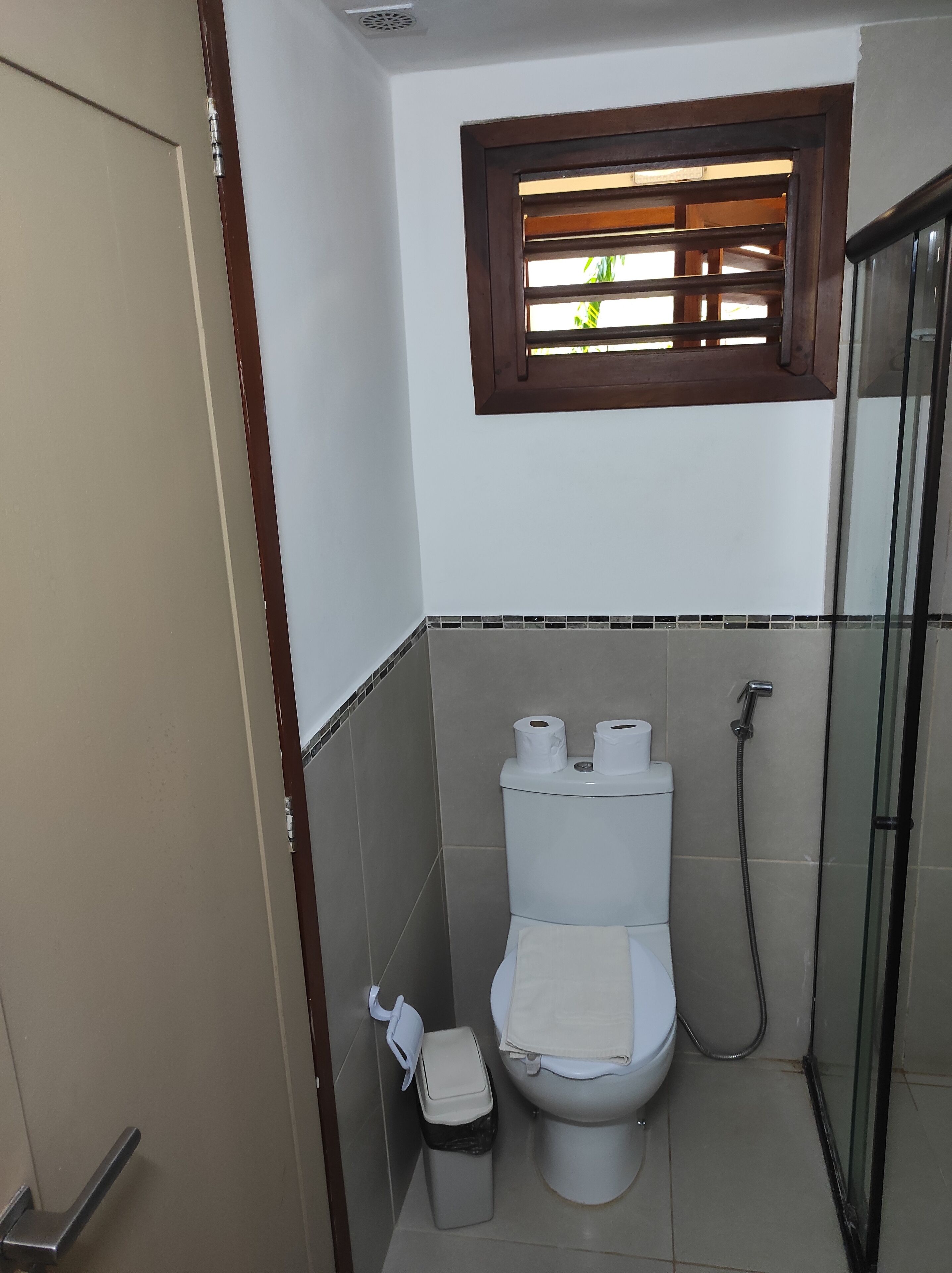 A small bathroom with a toilet, a trash bin, a shower, and a window with blinds.