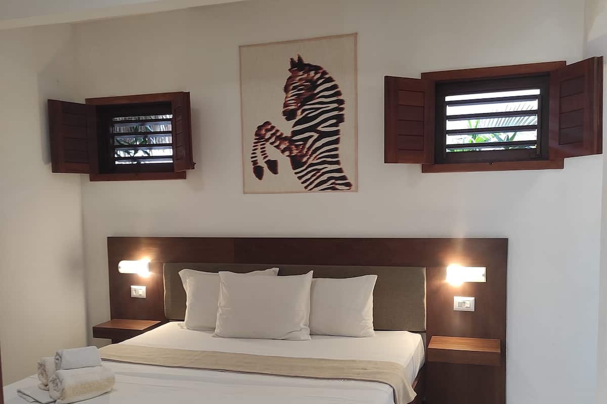 Un dormitorio con una cama grande, dos mesitas de noche con lámparas, una obra de arte con cebra en la pared y dos ventanas con contraventanas.