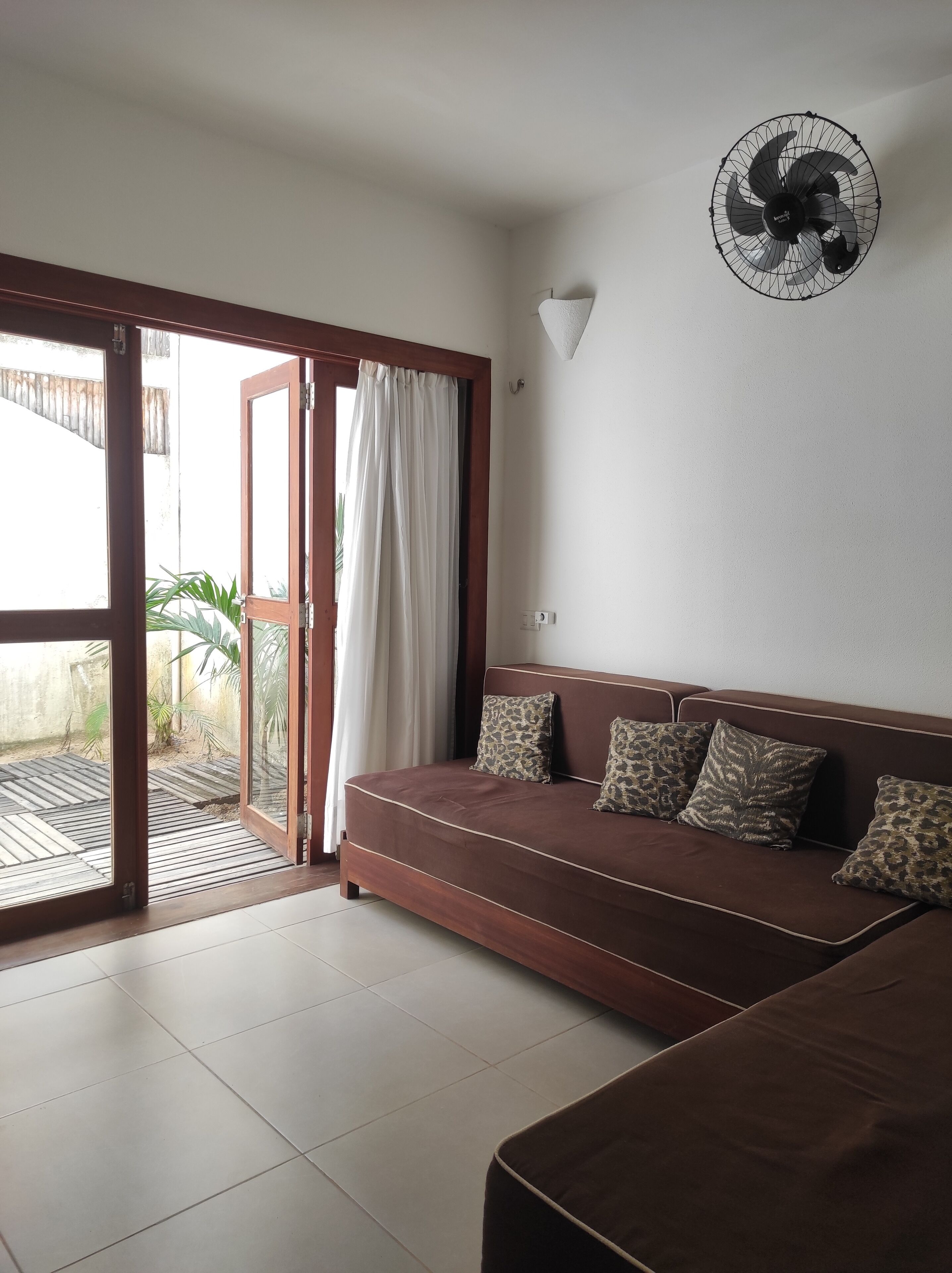 Ein Zimmer mit braunem Sofa, Deckenventilator und einer Glastür, die zu einem Außenbereich mit gefliestem Boden führt.
