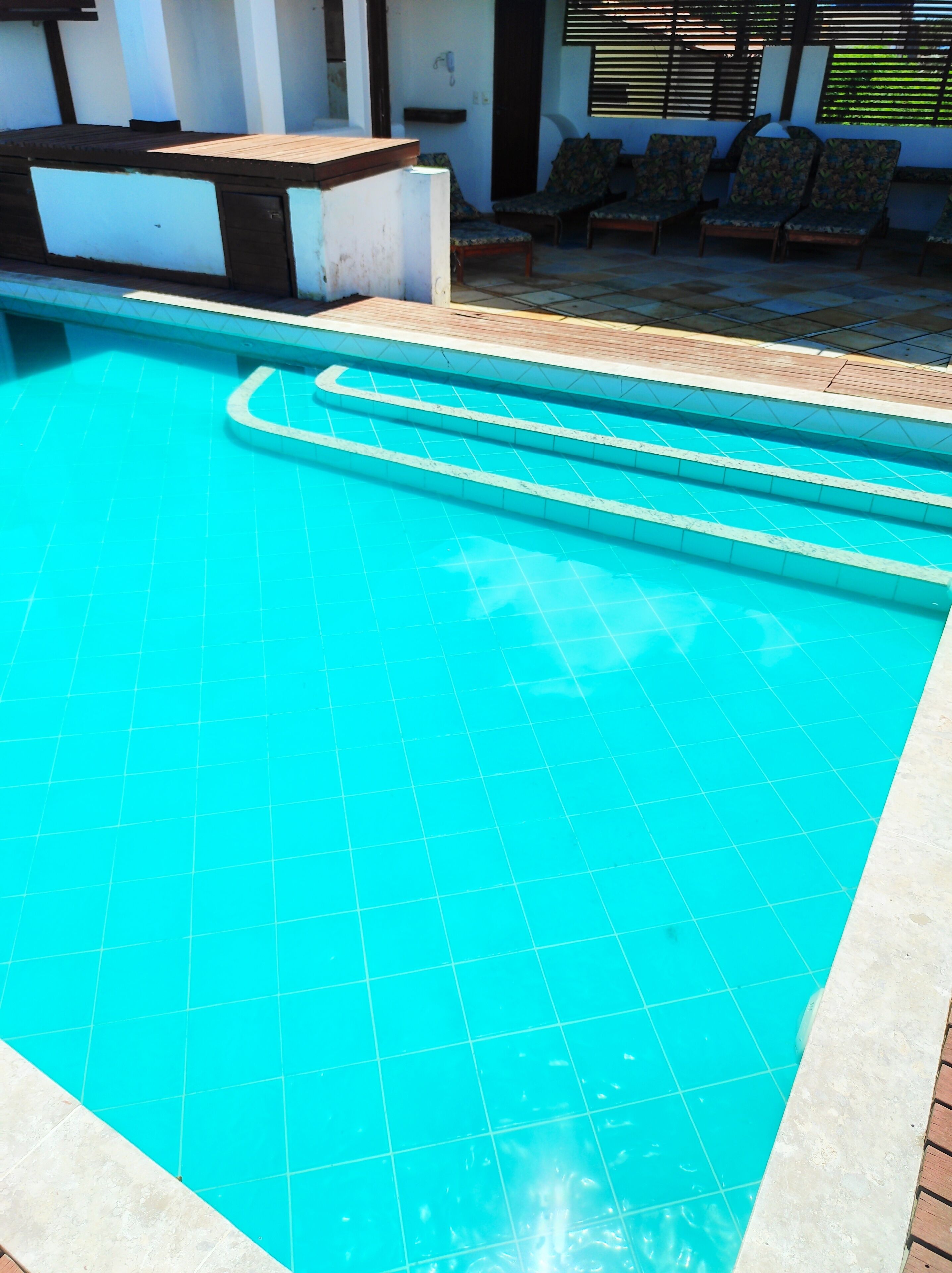 En swimmingpool med kristallklart blått vatten, kakelbeklädda trappsteg och ett trädäck med stolar.