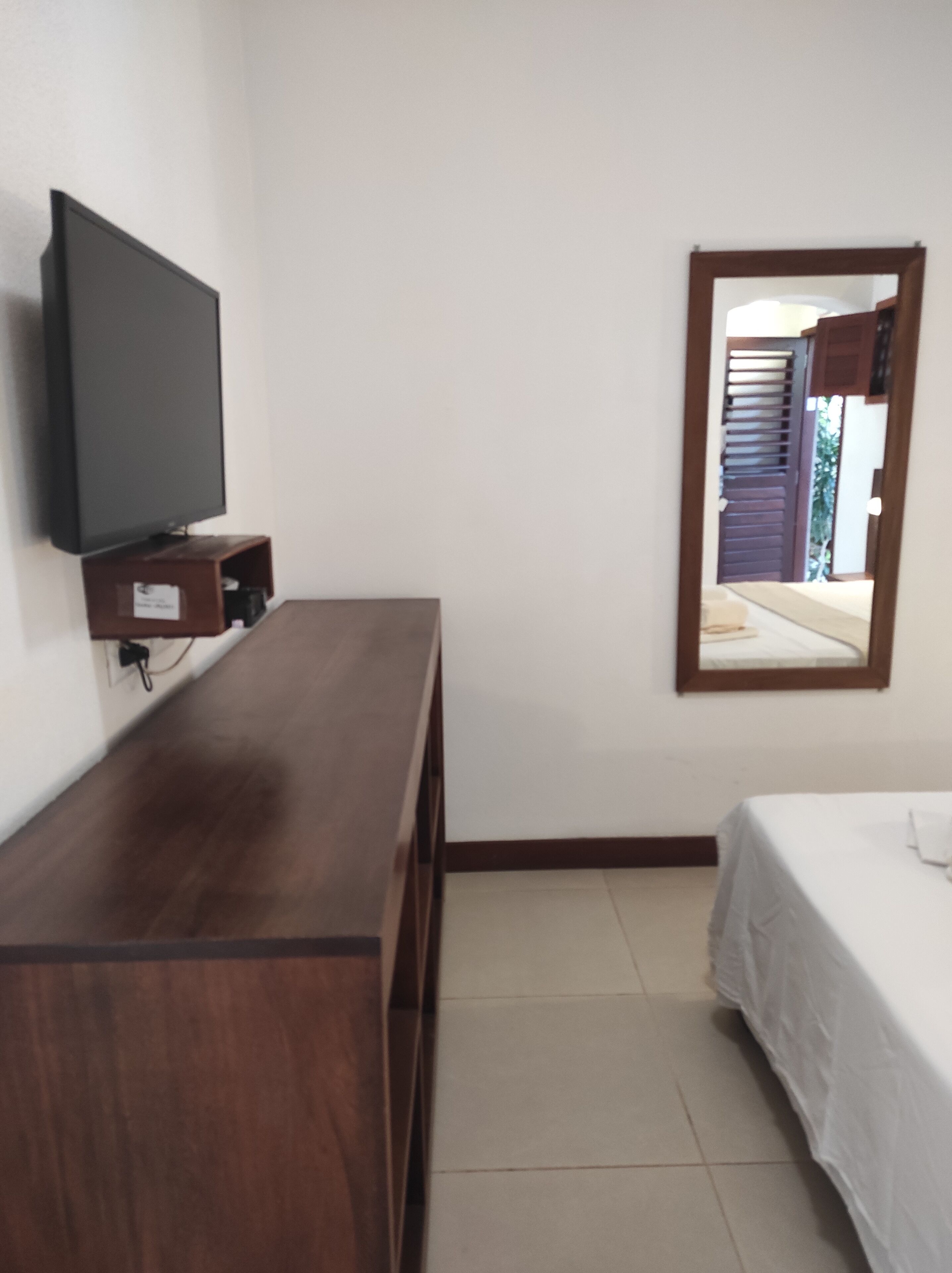 A bedroom with a wooden dresser, a television on a shelf, a bed with a white bedspread, and a mirror on the wall.